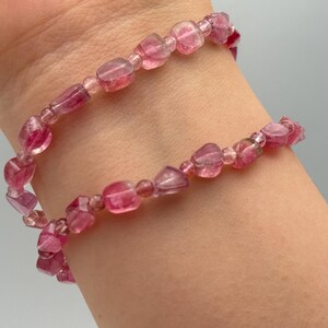 May include: A pink gemstone bracelet with a double strand design. The bracelet features small, irregular-shaped beads in varying shades of pink, strung together on a flexible cord. The beads appear translucent.