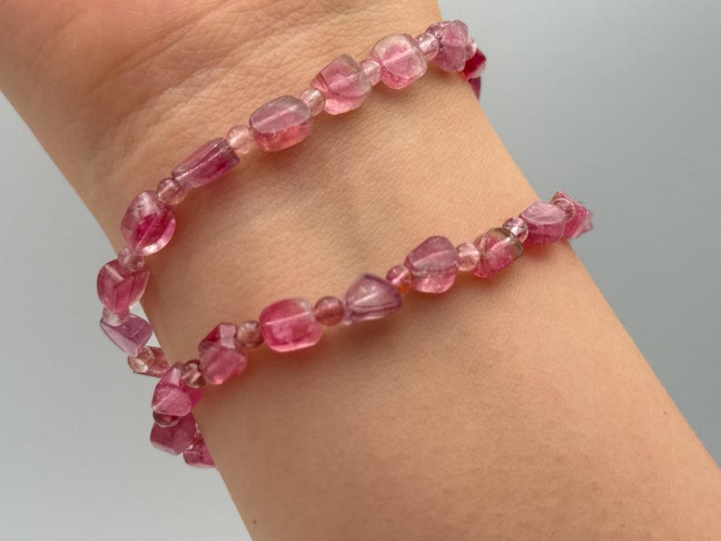 May include: A pink gemstone bracelet with a double strand design. The bracelet features small, irregular-shaped beads in varying shades of pink, strung together on a flexible cord. The beads appear translucent.