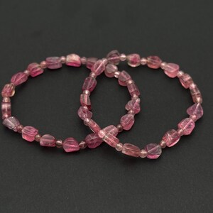 May include: Two pink tourmaline gemstone bracelets. Each bracelet is composed of irregular, faceted pink beads strung together. The beads vary in size and shade, creating a natural, organic look. The bracelets are displayed on a dark background.
