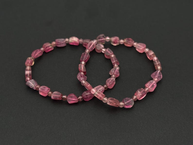 May include: Two pink tourmaline gemstone bracelets. Each bracelet is composed of irregular, faceted pink beads strung together. The beads vary in size and shade, creating a natural, organic look. The bracelets are displayed on a dark background.