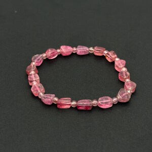 May include: A pink gemstone bracelet with a stretch design. The bracelet features a series of pink, rectangular-shaped beads, interspersed with smaller, round beads. The beads have a translucent quality, and the bracelet is set against a dark background.