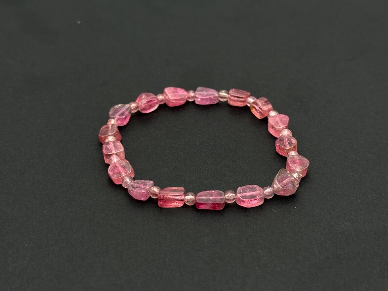 May include: A pink gemstone bracelet with a stretch design. The bracelet features a series of pink, rectangular-shaped beads, interspersed with smaller, round beads. The beads have a translucent quality, and the bracelet is set against a dark background.