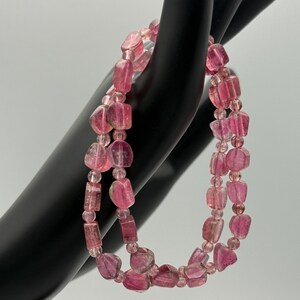 May include: A double-strand bracelet featuring pink tourmaline beads. The beads are a mix of irregular, faceted shapes and smaller, round beads. The bracelet is displayed against a black background, highlighting the pink hues of the gemstones. The bracelet is a piece of jewelry.