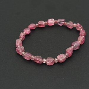 May include: A pink gemstone bracelet. The bracelet features a series of irregularly shaped, translucent pink beads strung together. The beads have a slightly rough texture and vary in size. The bracelet is displayed against a dark background.