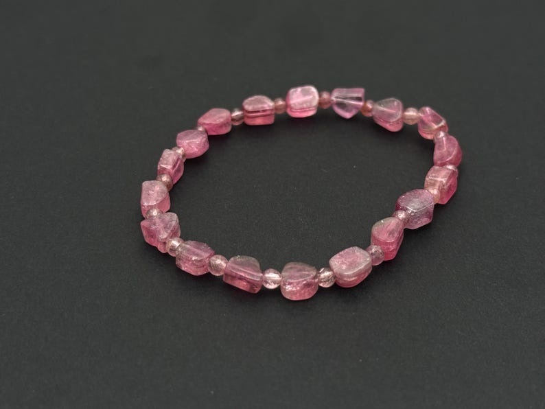 May include: A pink gemstone bracelet. The bracelet features a series of irregularly shaped, translucent pink beads strung together. The beads have a slightly rough texture and vary in size. The bracelet is displayed against a dark background.