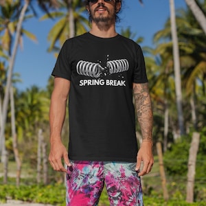 Puede incluir: Camiseta negra con un gráfico blanco de un muelle roto y las palabras "SPRING BREAK". La persona lleva pantalones cortos coloridos con un estampado floral tropical. Hay palmeras en el fondo.