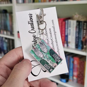 May include: A keychain featuring a miniature book with a green and pink cover and a pair of sneakers on the front. The book cover has the text "Buy My First Boy, First Love" on it. The keychain is attached to a silver key ring.