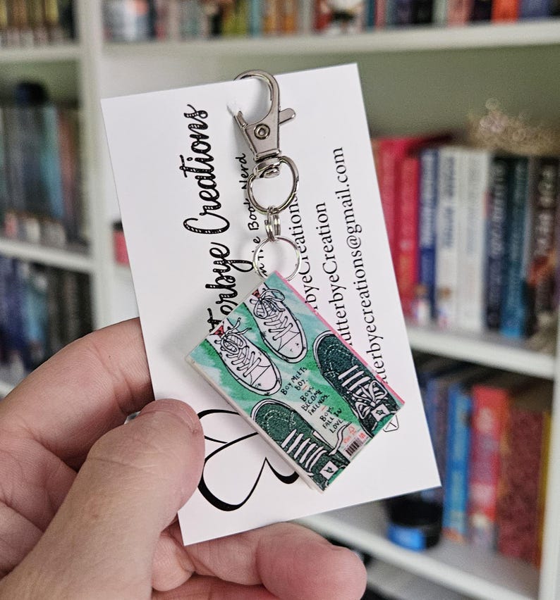 May include: A keychain featuring a miniature book with a green and pink cover and a pair of sneakers on the front. The book cover has the text "Buy My First Boy, First Love" on it. The keychain is attached to a silver key ring.