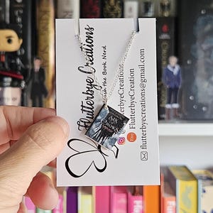May include: A silver necklace with a miniature book charm featuring the cover of "Six of Crows" by Leigh Bardugo. The book charm is dark blue and silver with a black and white illustration of a crow on the cover. The necklace is displayed on a white card with the Etsy logo and the text "Flutterbye Creations - with the Book Nerd".
