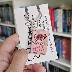 May include: A pink and white book charm keychain featuring the book cover of "Flawless" by Elsie Silver. The charm is attached to a silver key ring.
