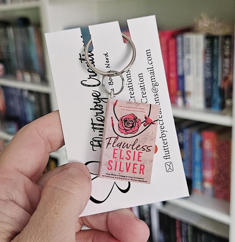May include: A pink and white book charm keychain featuring the book cover of "Flawless" by Elsie Silver. The charm is attached to a silver key ring.
