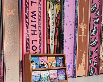 May include: A miniature wooden bookshelf displays several small books with colorful covers. The books are arranged in front of a row of larger books with pink and patterned spines. Titles include "The Love Hypothesis" and "Deep End".