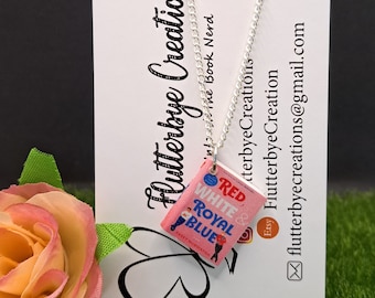 Può includere: Una collana con un ciondolo a forma di libro rosa e blu con il titolo "Red, White & Royal Blue" di Casey McQuiston. La collana è su una catena d'argento e pende da un biglietto bianco con il testo "Flutterbye Creations" e le informazioni di contatto.