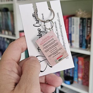 May include: A pink and white miniature book keychain with a silver key ring. The back of the book has text and a barcode. The keychain is held in a hand against a white card with black text that says "Etsy by the Nerd".