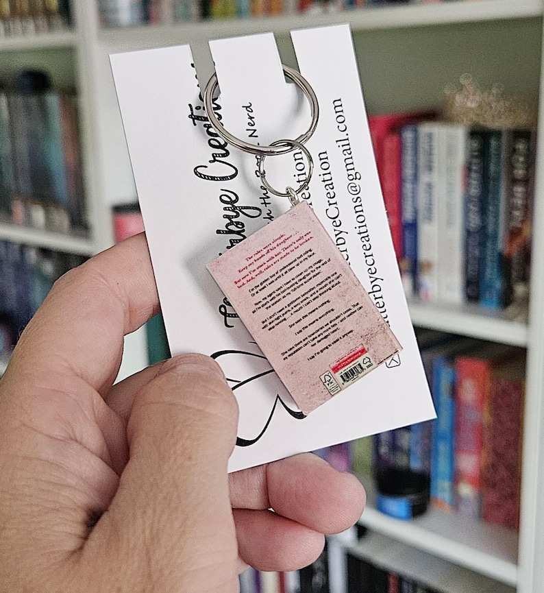 May include: A pink and white miniature book keychain with a silver key ring. The back of the book has text and a barcode. The keychain is held in a hand against a white card with black text that says "Etsy by the Nerd".