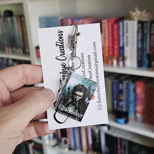 May include: A silver book charm keychain featuring a miniature book cover with the title "Quicksilver" and the author "Callie Hart". The book cover is blue and green with a silver design. The charm is attached to a silver chain and a silver lobster clasp.