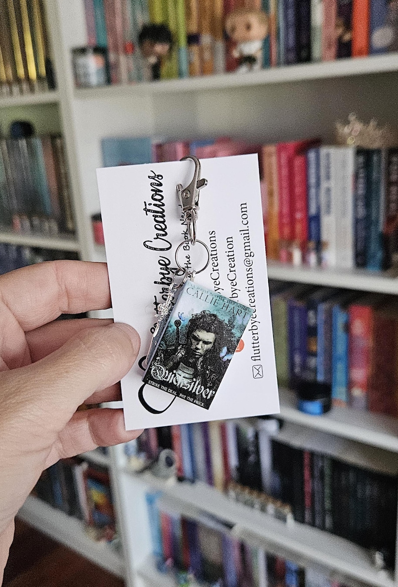 May include: A silver book charm keychain featuring a miniature book cover with the title "Quicksilver" and the author "Callie Hart". The book cover is blue and green with a silver design. The charm is attached to a silver chain and a silver lobster clasp.