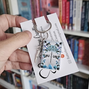 May include: A silver keychain featuring a miniature book with the title "Quick Silver" and a sword charm. The keychain is held in a hand against a background of bookshelves.