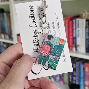 May include: A keychain featuring a miniature replica of the graphic novel "Heartstopper" by Alice Oseman. The keychain is made of resin and features a pink, blue, and green color scheme. The book cover features two characters, a boy and a girl, walking together. The keychain is attached to a silver metal key ring.