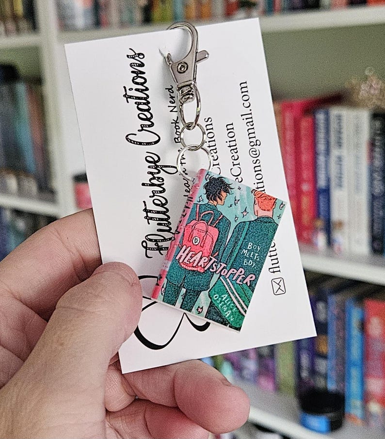 May include: A keychain featuring a miniature replica of the graphic novel "Heartstopper" by Alice Oseman. The keychain is made of resin and features a pink, blue, and green color scheme. The book cover features two characters, a boy and a girl, walking together. The keychain is attached to a silver metal key ring.