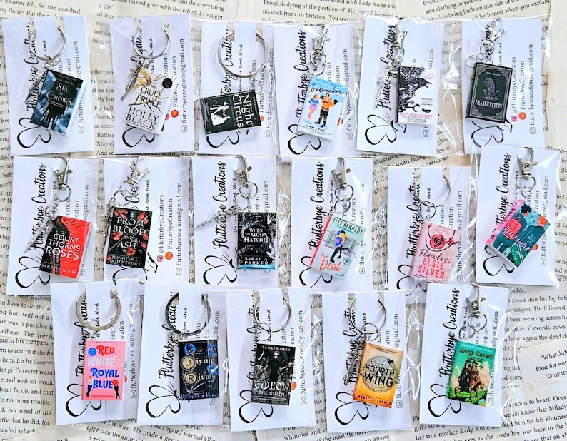 May include: A collection of 15 book-themed keychains featuring miniature book covers with titles such as "Cruel Prince", "Throne of Glass", "Red, White & Royal Blue", "The Deal", "Flawless, Elsie Silver", and "Fourth Wing". The keychains are displayed on a white background with a book page pattern.