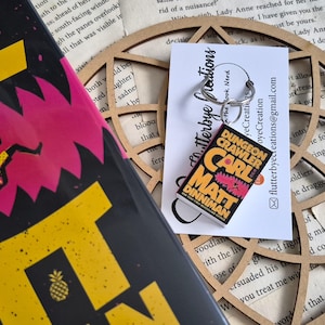 May include: A miniature book keychain featuring the cover of Dungeon Crawler Carl by Matt Dinniman, with a black, yellow, and pink design. The keychain is attached to a silver metal ring and displayed on a white card with black text.