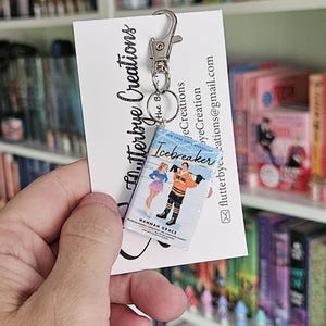 May include: A keychain featuring a miniature paperback book with the title "Icebreaker" by Hannah Grace. The book is blue with white text and a colorful illustration of two people ice skating. The keychain is attached to a silver key ring.