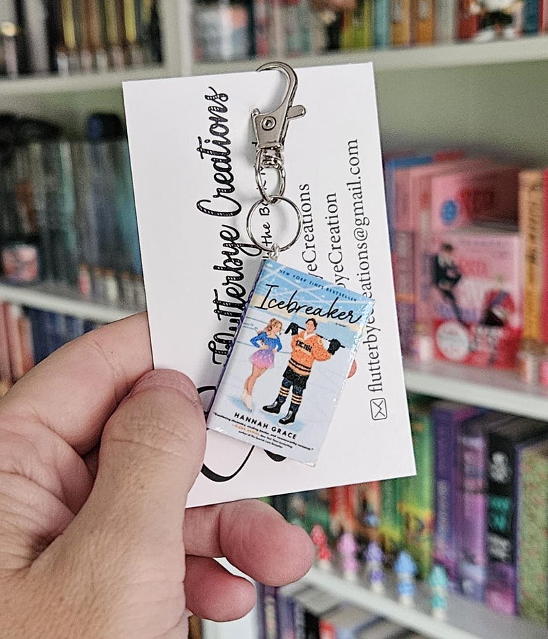 May include: A keychain featuring a miniature paperback book with the title "Icebreaker" by Hannah Grace. The book is blue with white text and a colorful illustration of two people ice skating. The keychain is attached to a silver key ring.