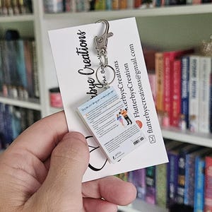May include: A miniature book keychain with a silver key chain. The book is a paperback with a white cover and a colorful image of a couple on the back. The text on the back of the book reads "Only the one thing you can't control is how you choose to react to it."