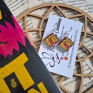 May include: A pair of miniature book earrings featuring the cover art for Dungeon Crawler Carl by Matt Dinniman. The earrings are attached to a white display card with black text and a butterfly logo, positioned next to a book and a wooden cutout.