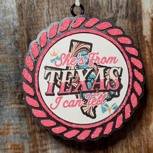May include: A round hanging ornament featuring a black scalloped border with pink glitter rope detailing. The center displays a white background with a map of Texas, a small crown, and the text "She's From TEXAS I can Tell" in pink and black.