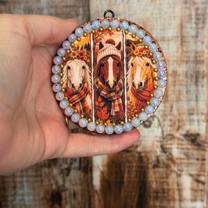 May include: A circular wooden ornament featuring three panels of horses in autumn attire, including scarves and hats, surrounded by a border of iridescent white beads and small gold metallic spheres, with a black metal loop at the top.