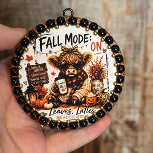 Può includere: Un ornamento decorativo rotondo con una mucca highland che indossa occhiali da sole e una fascia leopardata, mentre tiene una tazza di caffè. Il testo recita FALL MODE: ON e Leaves, Lattes AND A LITTLE ATTITUDE. Il bordo è rifinito con perline nere e dorate.