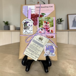 May include: A wrapped gift package with a variety of items, including a pen, stickers, and a tea packet. The package is tied with purple and pink twine. The stickers read "My Weekend Is All Booked" and "Midnight Readers Book Club".