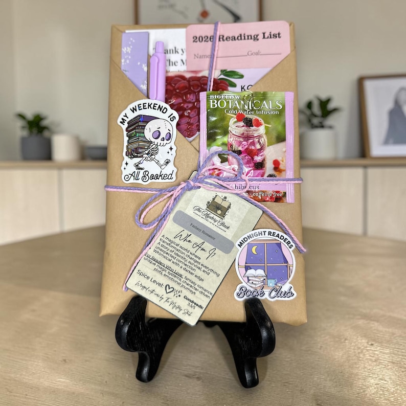 May include: A wrapped gift package with a variety of items, including a pen, stickers, and a tea packet. The package is tied with purple and pink twine. The stickers read "My Weekend Is All Booked" and "Midnight Readers Book Club".