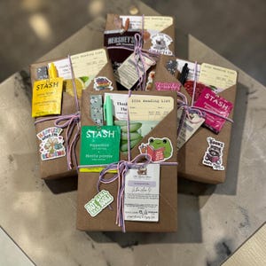 May include: Several brown paper packages tied with purple and white string. Each package has stickers, tea, and a Hershey's chocolate bar. Some packages have the text "2026 Reading List" and "Happiest when I'm READING".