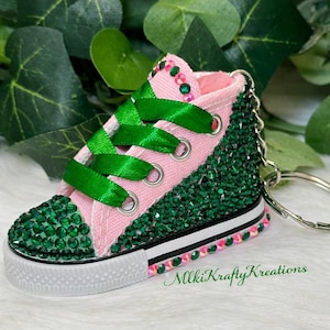 May include: A miniature pink high-top sneaker keychain, embellished with green rhinestones and green ribbon laces. The shoe has a white sole with a black stripe and is accented with pink and green rhinestones. The text "MlkiKraftyKreations" is visible.