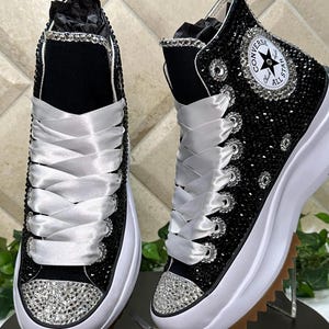 May include: A pair of black high-top sneakers featuring a thick white platform sole with a brown serrated tread. The upper is covered in black crystals, with silver crystal-encrusted toe caps, wide white satin ribbon laces, and a circular logo patch.
