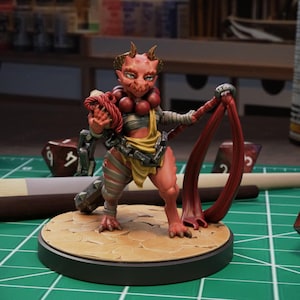 May include: A miniature fantasy figure of a red-skinned demon with horns, holding a red rope. The figure is on a round base, surrounded by dice and a paintbrush on a green surface. The figure is painted with detailed shading.