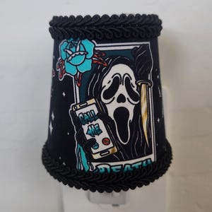 Puede incluir: Luz de noche negra con una pantalla de tela decorativa con un gráfico de máscara de Scream. El diseño incluye una rosa turquesa, un cuchillo y el texto "CALL ME DEATH". La pantalla está adornada con una cuerda negra.