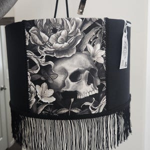 Puede incluir: Pantalla de lámpara negra con estampado floral y calavera. La pantalla tiene flecos negros en la parte inferior y un ribete negro. El estampado presenta una calavera rodeada de flores y follaje en tonos grises y blancos.