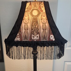 May include: Black floor lamp with a decorative lampshade featuring a gothic-style design. The lampshade has a black fringe and a patterned design with figures and a central moon. The lamp is a decorative home decor item.