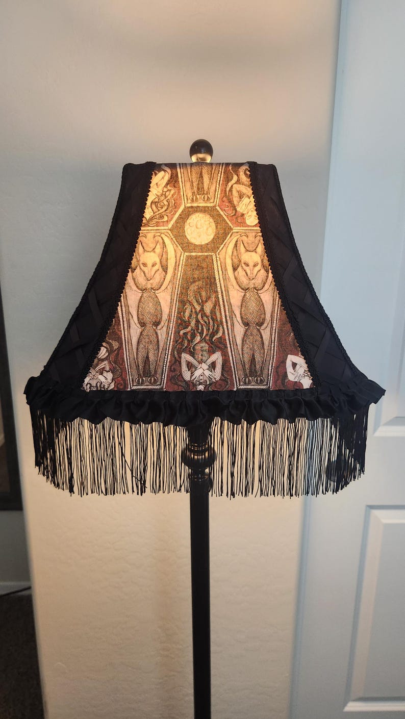 May include: Black floor lamp with a decorative lampshade featuring a gothic-style design. The lampshade has a black fringe and a patterned design with figures and a central moon. The lamp is a decorative home decor item.