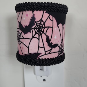 May include: A decorative night light with a pink and black Halloween theme. The cylindrical shade features a pink background with black bats, spiderwebs, and stars. The top and bottom edges are trimmed with black decorative trim.