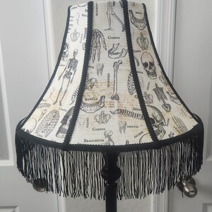 May include: A bell-shaped lamp shade featuring a cream-colored fabric with black anatomical illustrations of human skeletons, skulls, and bones, labeled with Latin text. The shade has black braided trim and a long black fringe along the bottom edge.