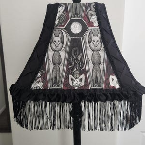 May include: Black lamp with a decorative lampshade featuring a gothic design. The lampshade has a black fringe and a detailed illustration of bats, coffins, and figures. The lamp is a unique home decor item.
