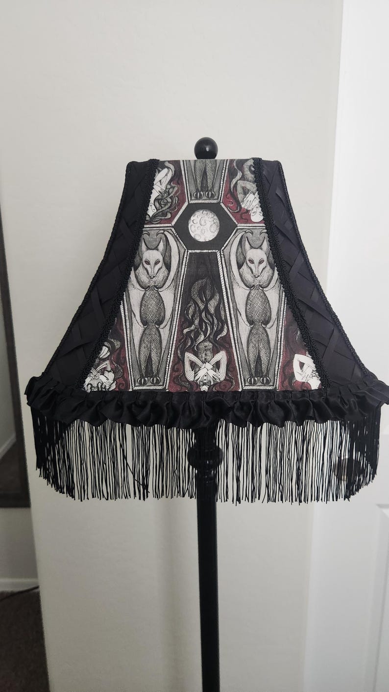 May include: Black lamp with a decorative lampshade featuring a gothic design. The lampshade has a black fringe and a detailed illustration of bats, coffins, and figures. The lamp is a unique home decor item.