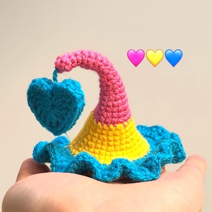 May include: A handmade crochet decoration featuring a blue heart suspended from a pink and yellow hat-like structure with a blue ruffled base. The design incorporates the colors of the Pansexual Pride flag. The background is a solid, neutral color.