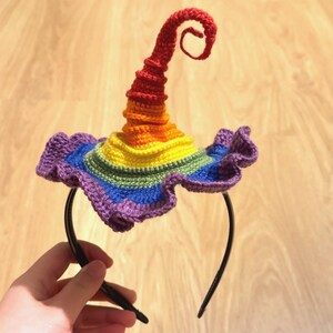 May include: A black plastic headband topped with a crocheted witch hat featuring a spiraled, pointed crown and a ruffled brim. The hat displays a vibrant rainbow color gradient, transitioning from purple at the base to blue, green, yellow, orange, and red at the tip.