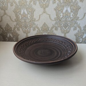 May include: A dark brown, handcrafted ceramic plate with a decorative, carved floral border. The plate has a wide, shallow design and a smooth, polished surface. The plate is likely used for serving food.